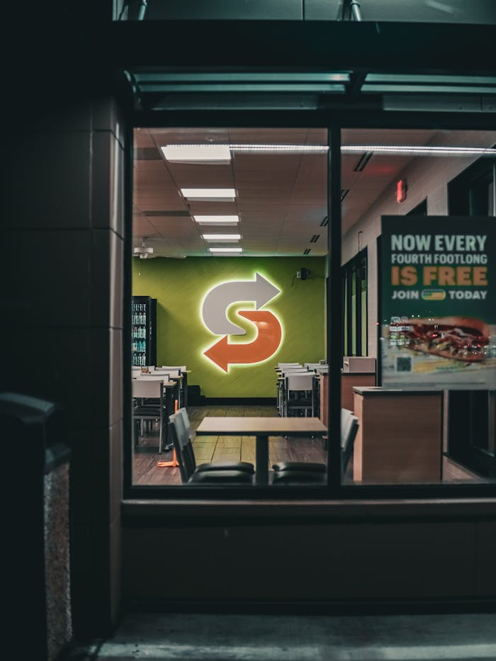 A dimly lit restaurant interior with a glowing neon logo sign, viewed from outside at night.